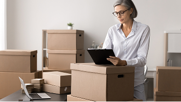 A business co-owner taking inventory in a storage room filled with cardboard boxes, using a clipboard and laptop